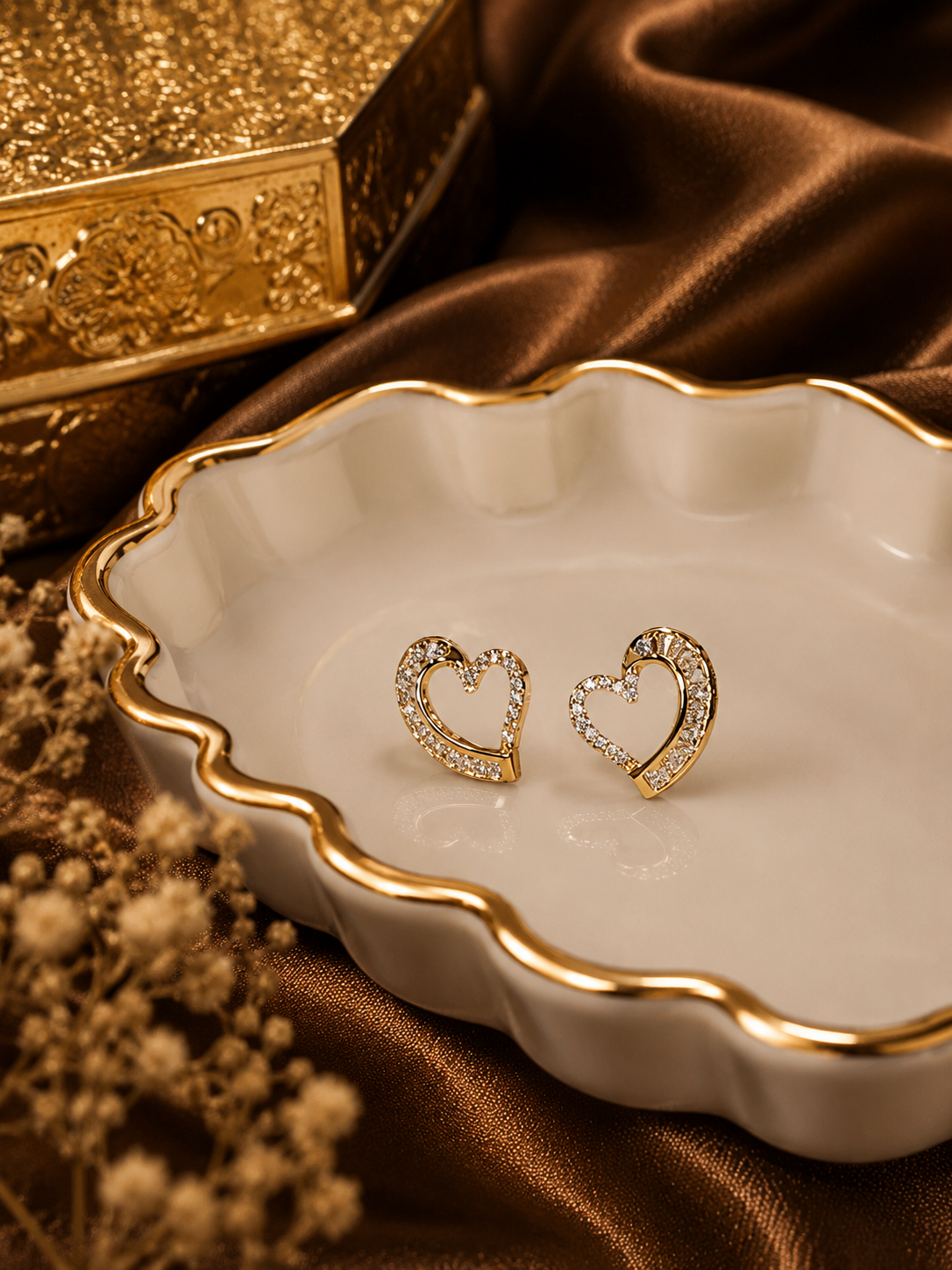 Heart-shaped anti tarnish earrings on a white dish with a gold box and brown fabric in the background