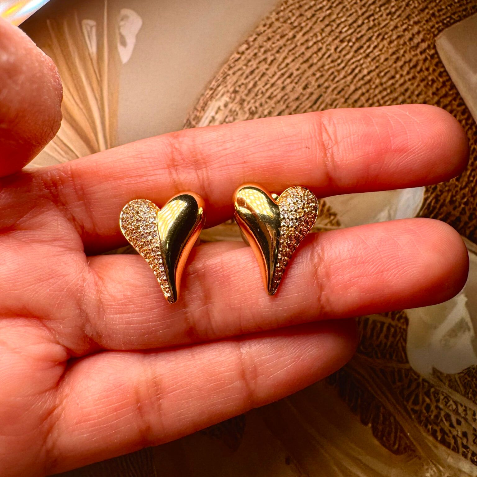 Gold heart-shaped earrings held between fingers against a patterned background