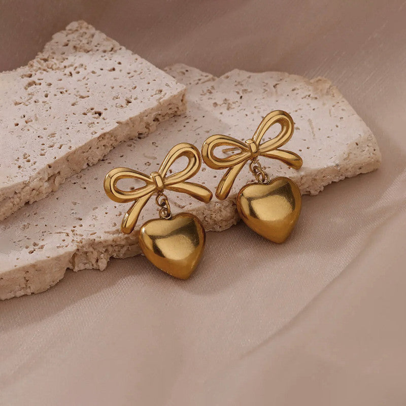 Gold bow and heart-shaped earrings on a textured beige surface