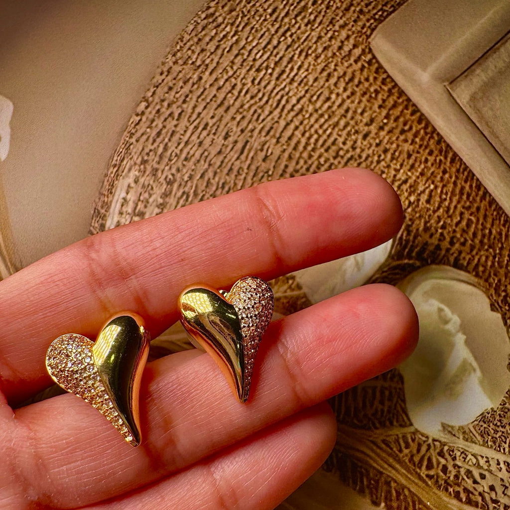 Gold heart-shaped earrings held between fingers against a textured beige background