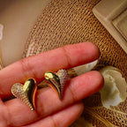 Gold heart-shaped earrings held between fingers against a textured beige background