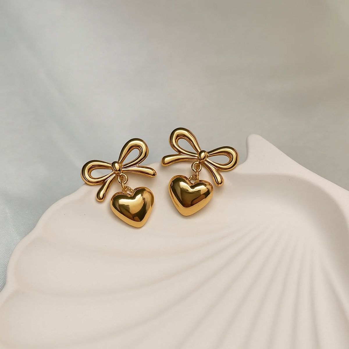 Gold bow and heart earrings on a shell-shaped stand with a neutral background
