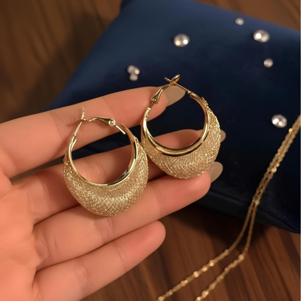 Gold hoop earrings held in a hand with a dark blue fabric background.