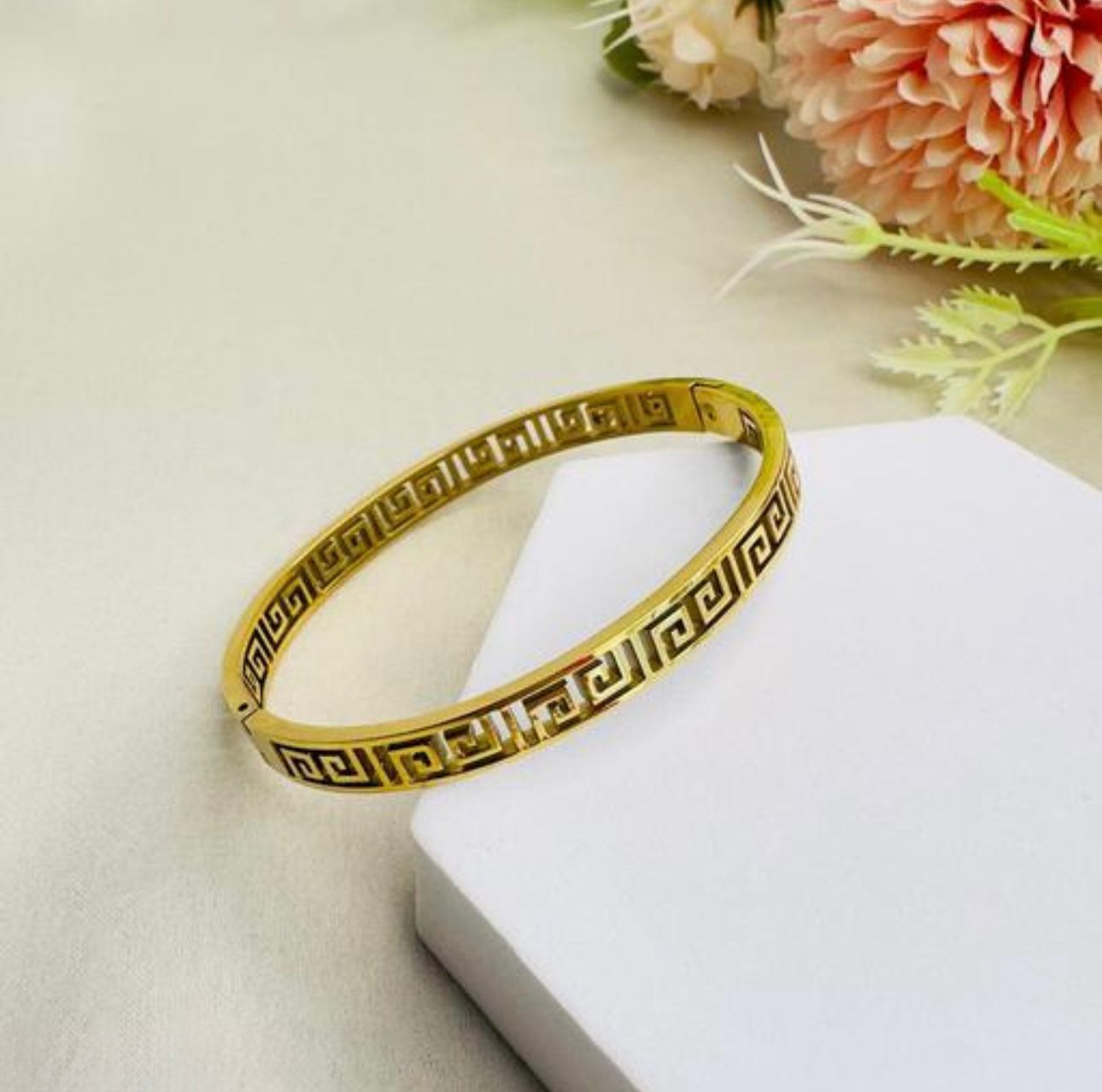 Versace Inspired Bangle - Valerio Gold bracelet on a white surface with flowers in the background